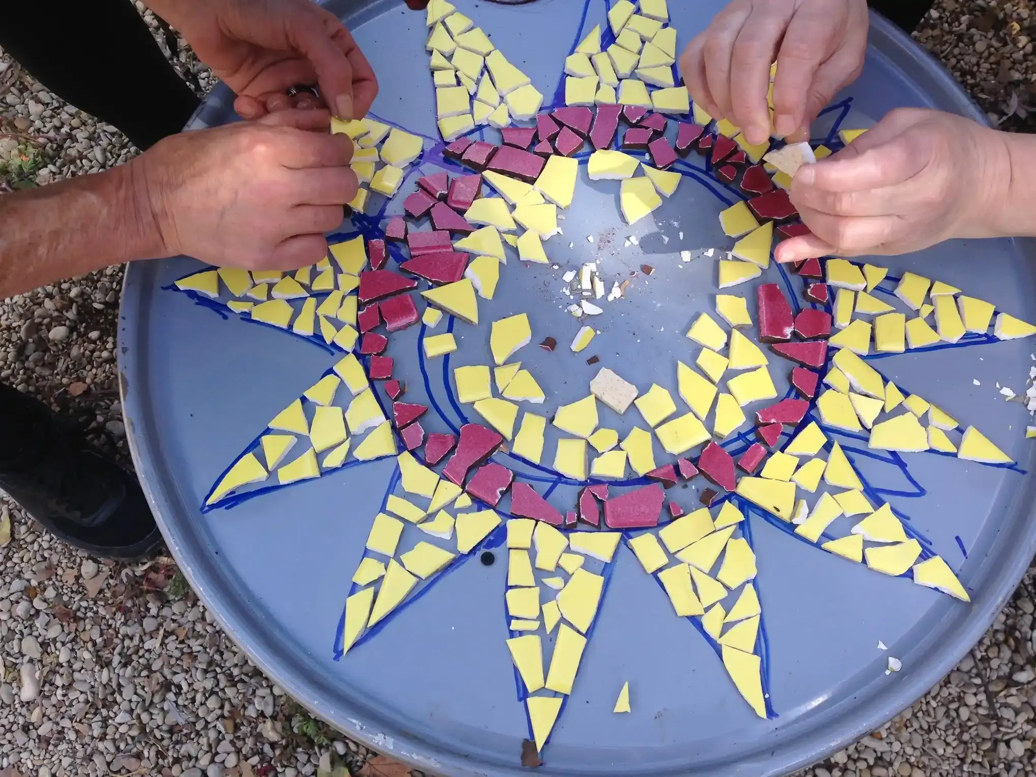 Des mains posent des tesselles jaunes et magenta sur un soleil en mosaïque