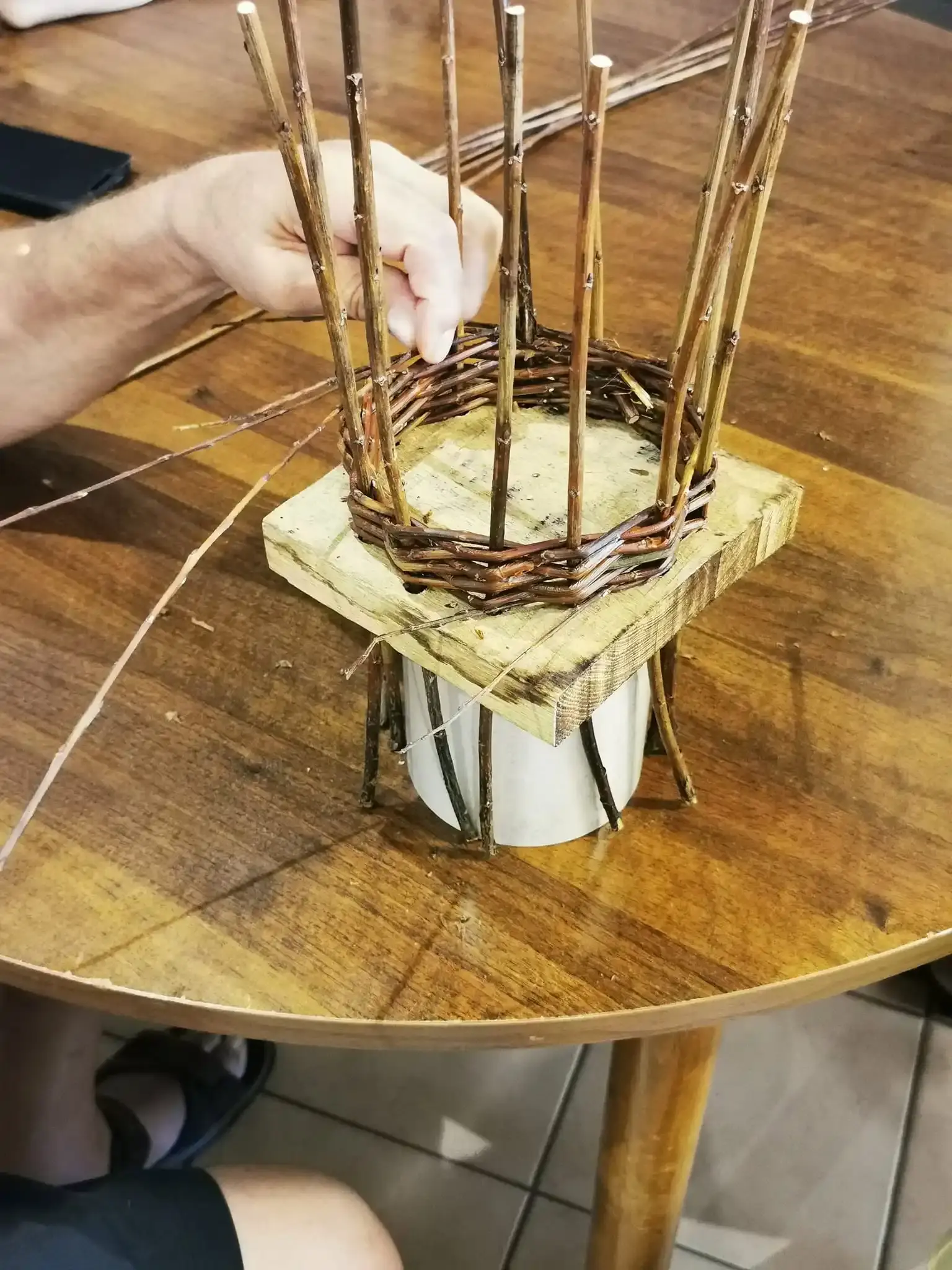 Des mains tressent des brins d’osier autour d’une petite structure en bois